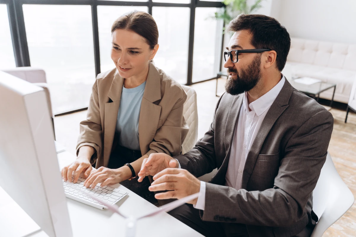 Two business people collaborating on a project, using a desktop computer in a bright, modern office environment