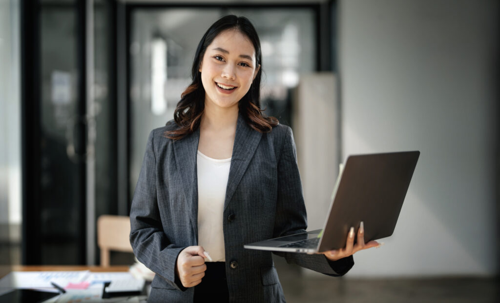 A young business professional holding a laptop while in a modern office setting.