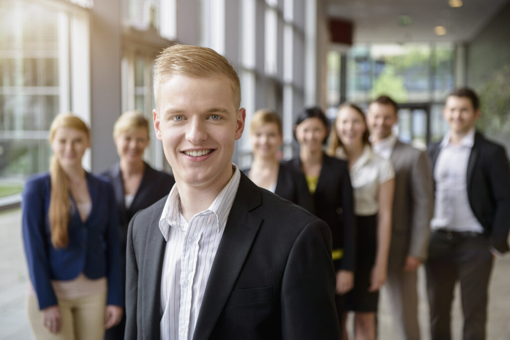 A young professional standing confidently in front of his peers in a modern corporate environment